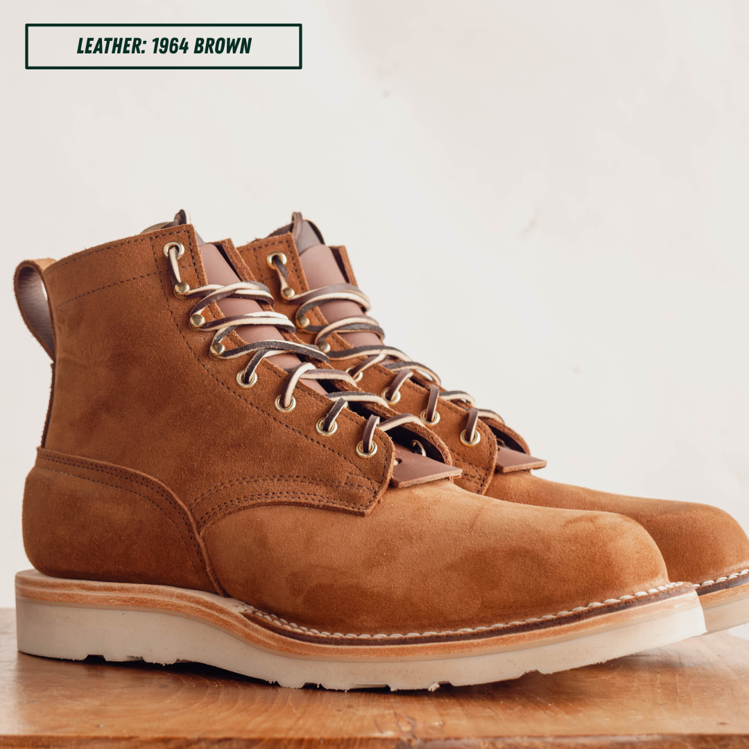 A pair of brown suede lace-up work boots with metal eyelets and a thick white sole, displayed on a wooden surface against a light background. A text label in the top left reads 'LEATHER: 1964 BROWN'.