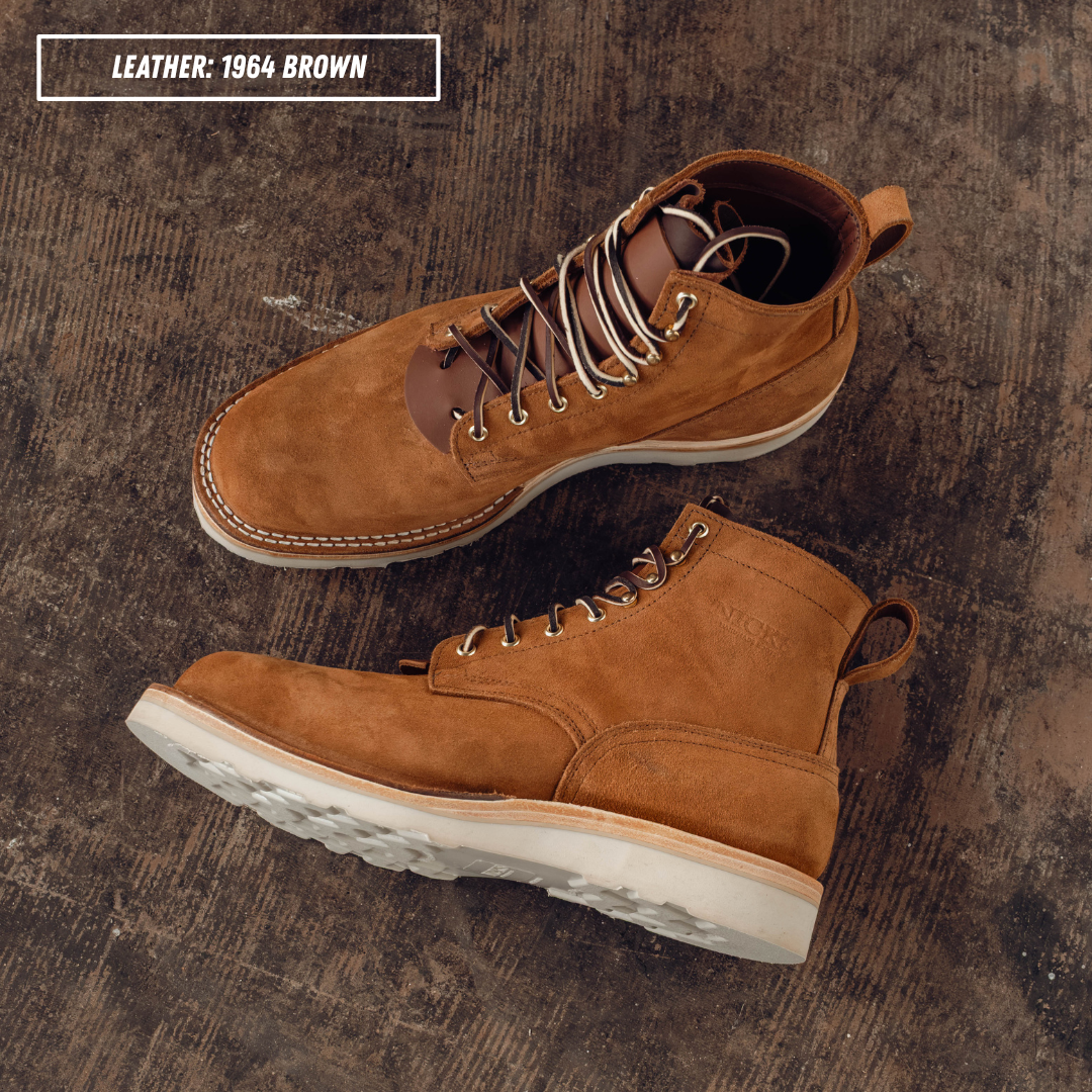 A pair of brown leather lace-up boots arranged on a dark textured concrete surface, with a text label reading 'LEATHER: 1964 BROWN' in the top left corner.