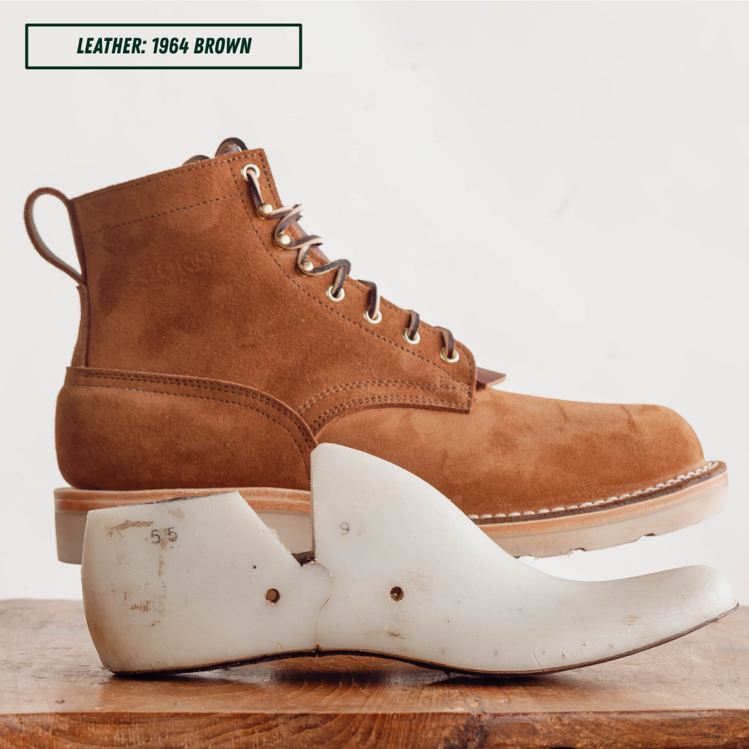 A brown suede lace-up boot shown in profile with a white wooden shoe tree in front, on a wooden surface against a light background. A text box at the top reads 'LEATHER: 1964 BROWN'.