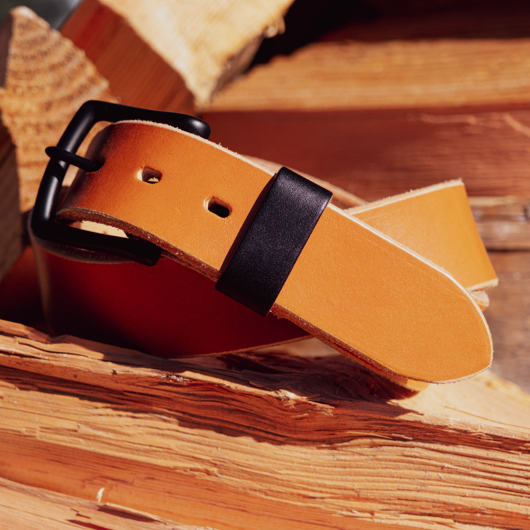 A close-up of a tan leather belt with a black buckle and black keeper loop, resting on rustic wood logs.