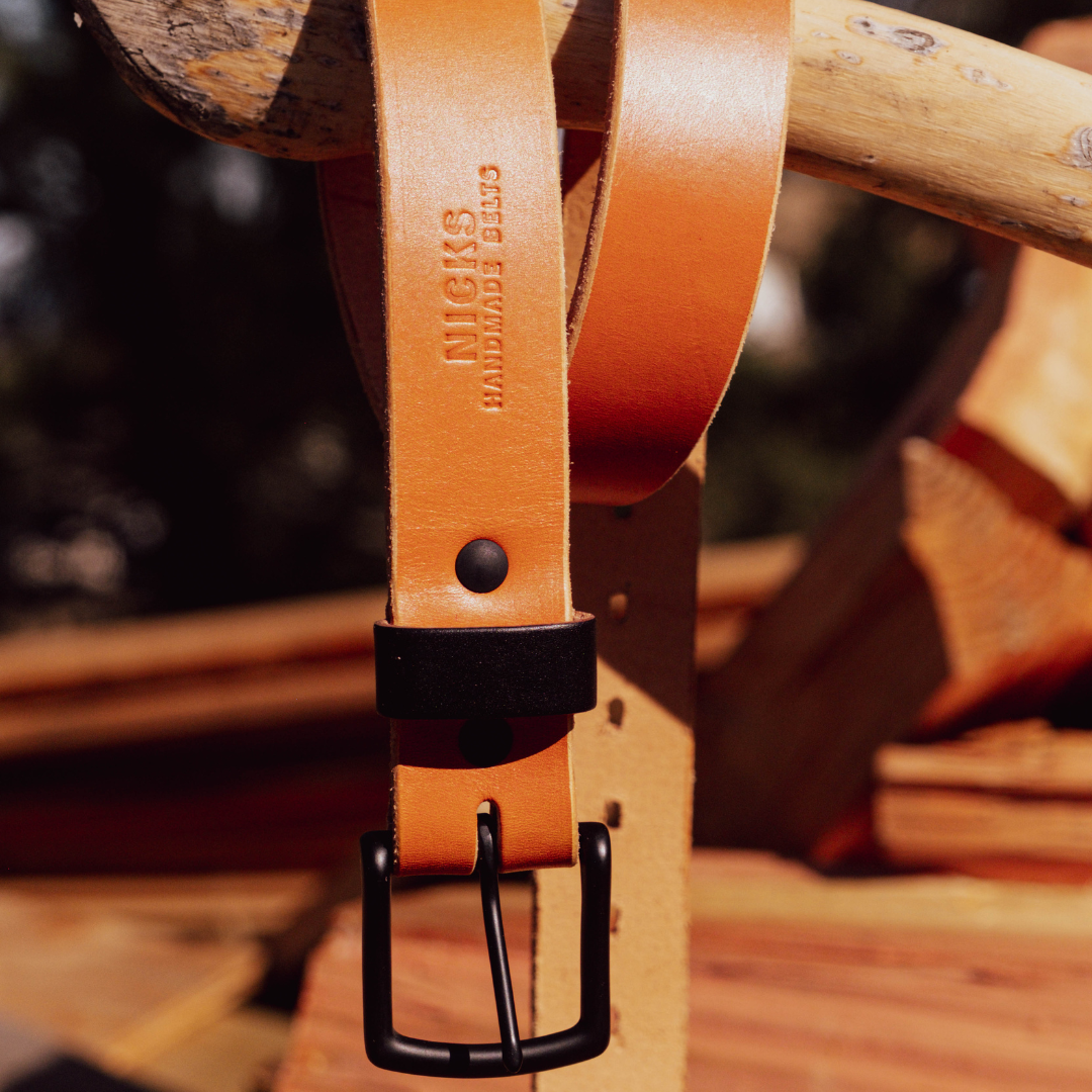 A close-up of a tan leather belt with a black buckle and black keeper, hanging from a rustic wooden branch. The brand name 'NICKS HANDMADE BELTS' is embossed on the leather.