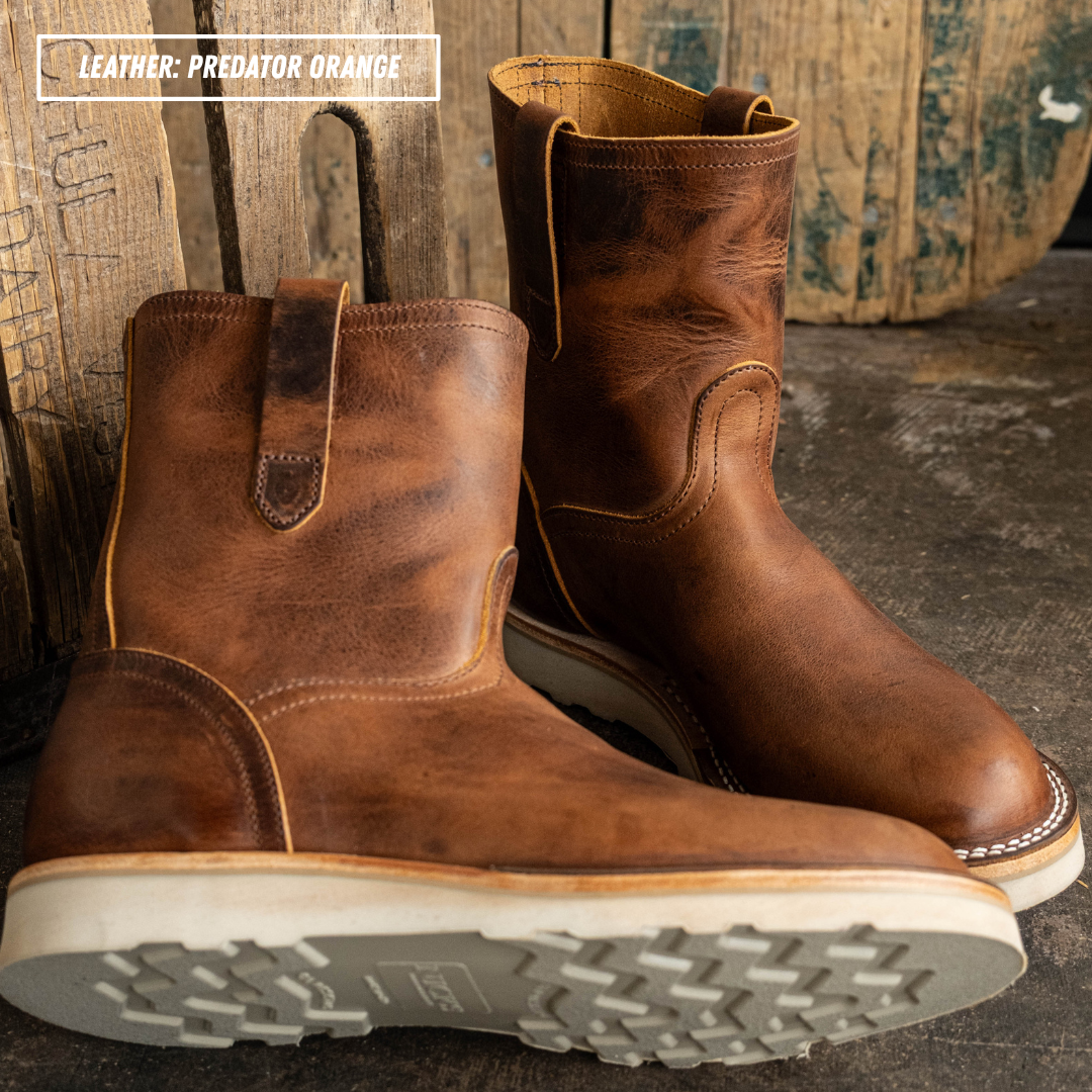 Brown leather pull on boots with white outsole laying on textured concrete and wooden pallet wall in the background.