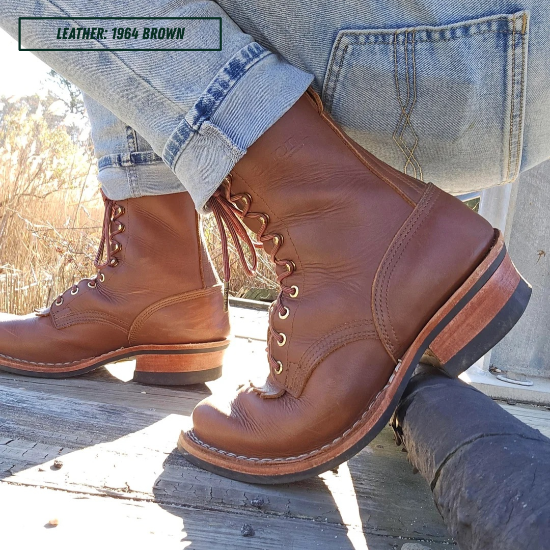 A person wearing a pair of brown leather lace-up boots with light blue jeans, sitting outdoors on a weathered wooden surface. A text overlay in the top left reads 'LEATHER - 1964 BROWN'.