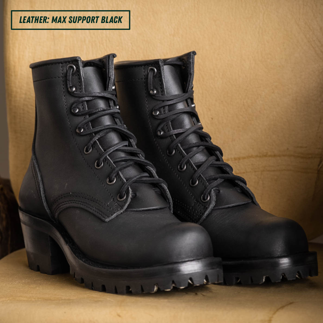 A pair of black leather lace-up boots with chunky lug soles and low block heels, displayed on a tan textured surface against a matching tan background. A text label in the top-left corner reads 'LEATHER: MAX SUPPORT BLACK'.