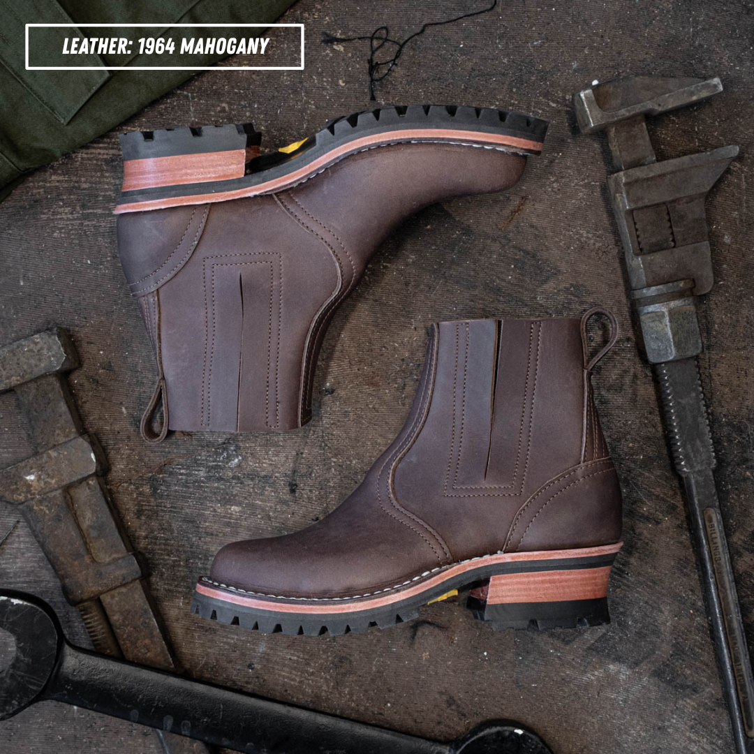 Two dark brown leather Chelsea boots with elastic side panels and lug soles, arranged on a rustic wooden surface with metal tools. A text label in the top left reads 'LEATHER: 1964 MAHOGANY'.