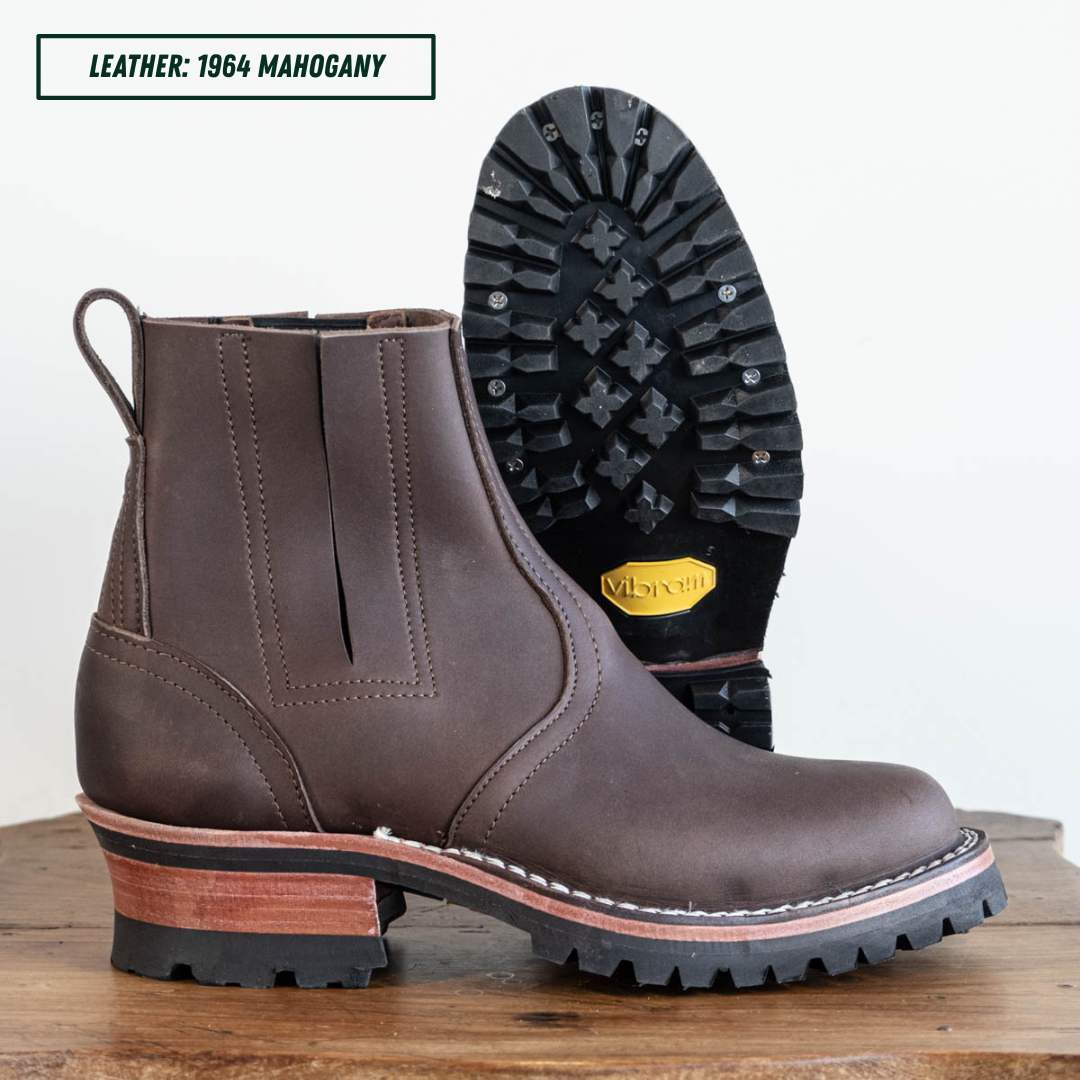 A dark brown leather ankle boot with a chunky block heel, shown in profile with its treaded sole facing up. The boot is displayed on a wooden surface against a plain white background, with a text label in the top left corner reading 'LEATHER: 1964 MAHOGANY'.