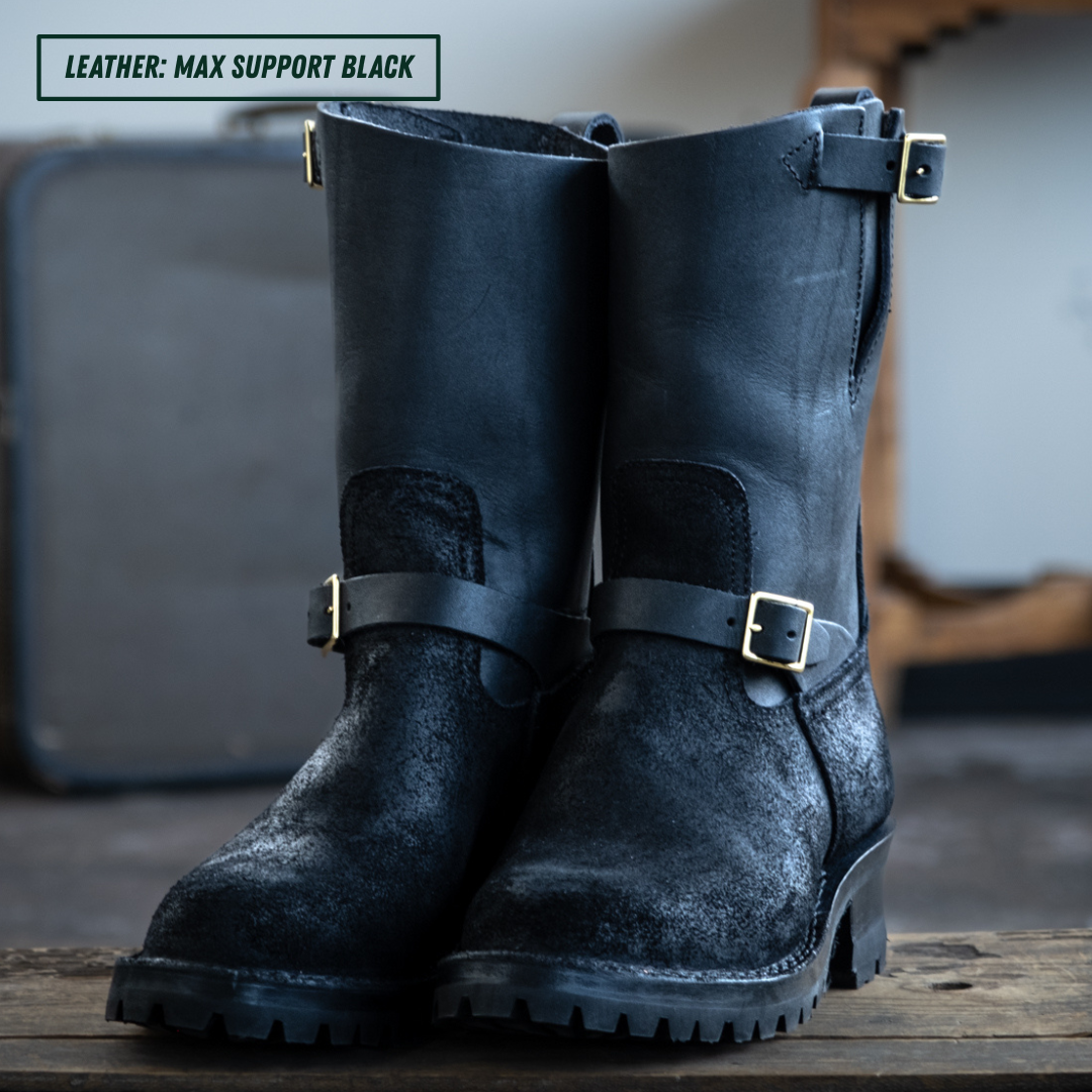 A pair of tall black leather boots with distressed textures and brass buckle straps, displayed on a wooden surface with a blurred background.