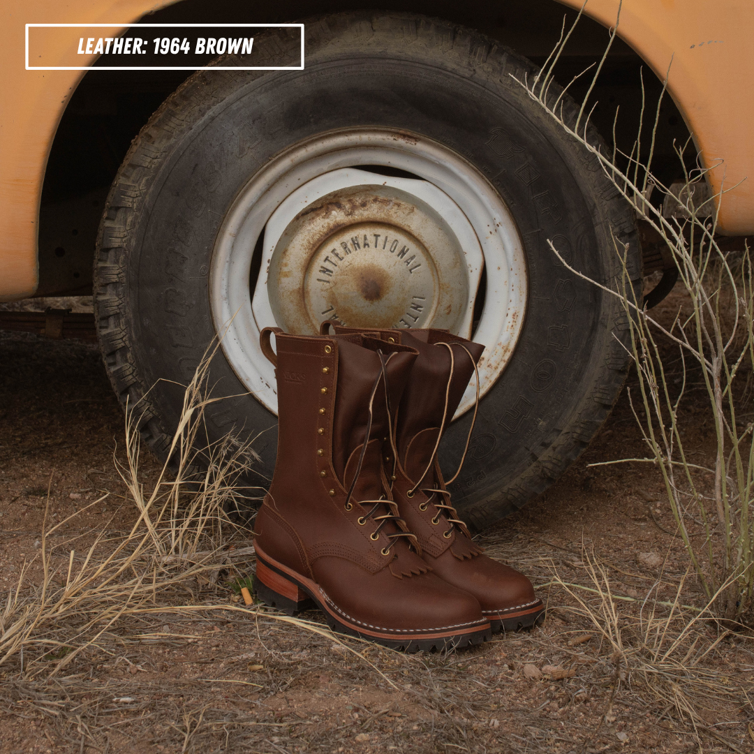 A pair of 1964-style brown leather lace-up work boots placed in front of a vintage vehicle wheel on dry grass and dirt ground. A text label in the top-left reads 'LEATHER: 1964 BROWN'.