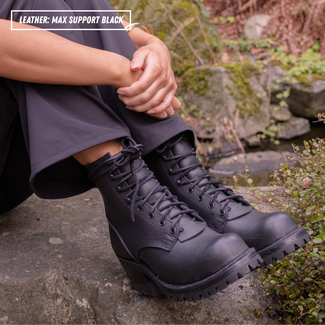 A person seated outdoors on rocks wearing black lace-up combat-style boots with a chunky sole. The photo includes a text overlay in the top left that reads 'LEATHER: MAX SUPPORT BLACK'.