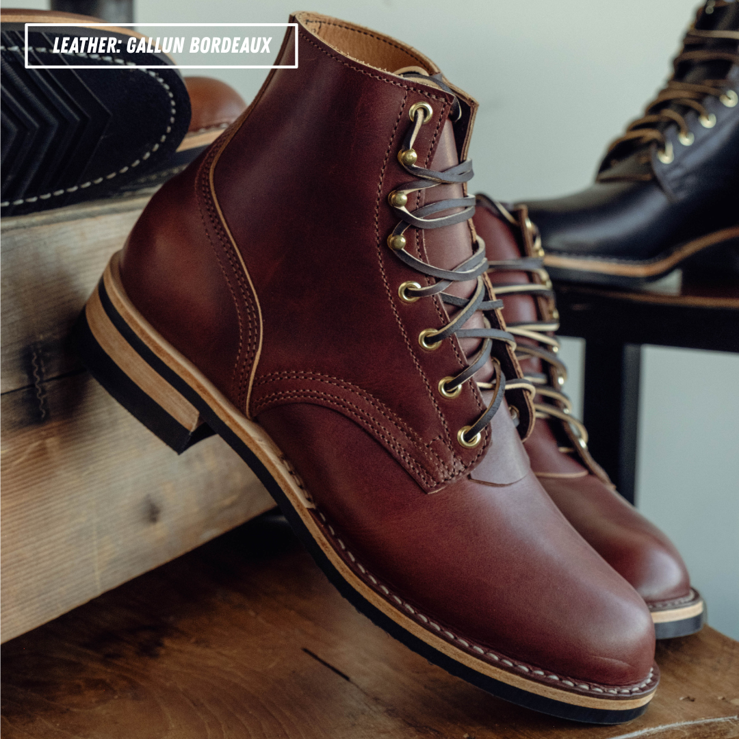 A pair of dark reddish-brown leather lace-up ankle boots with tan stitching and metal eyelets, displayed on a wooden surface with other boots in the background.