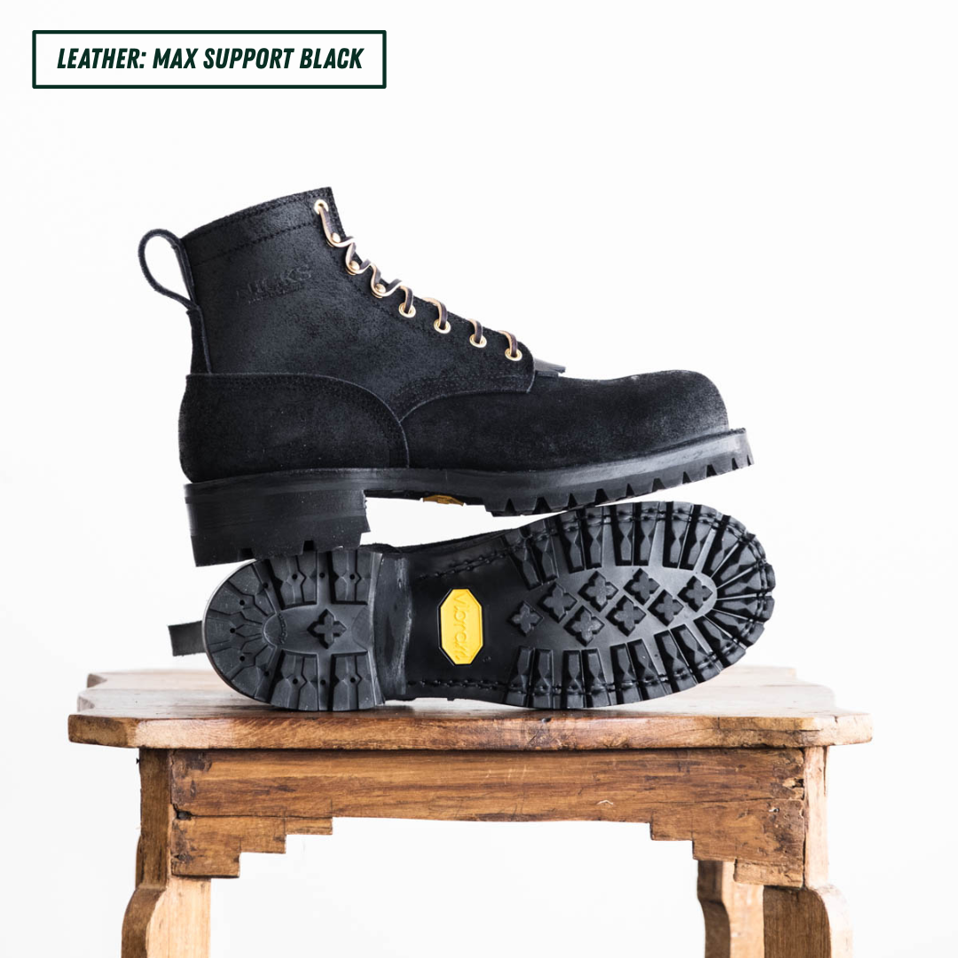 All black leather boots with lug soles. Boots sitting on wooden table, one laying down showing outsole and the other sitting side profile on top. Top left reads "LEATHER: MAX SUPPORT BLACK."