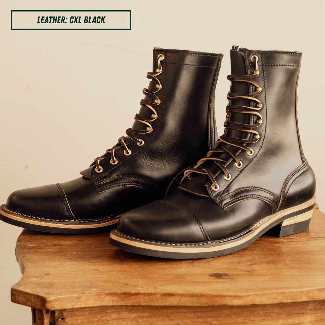 A pair of black leather lace-up boots with gold-toned metal eyelets and a light tan sole edge, displayed on a rustic wooden table against a warm beige background. A text overlay in the top left reads 'LEATHER: CXL BLACK'.