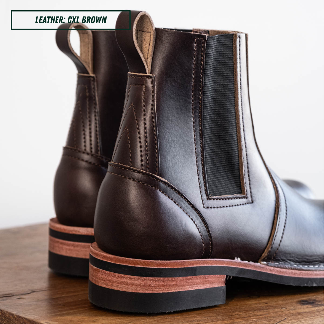 A pair of dark brown Chelsea boots made of cxl brown leather, shown from a rear three-quarter angle on a wooden surface against a white background. A text overlay in the top left corner reads 'LEATHER: CXL BROWN'.