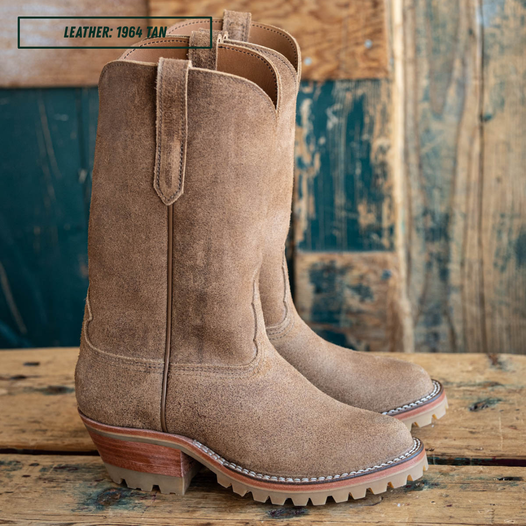 A pair of tan suede western-style boots shown in side profile on a wooden table with a wood pallet background. A small label in the top left reads 'LEATHER: 1964 TAN'.