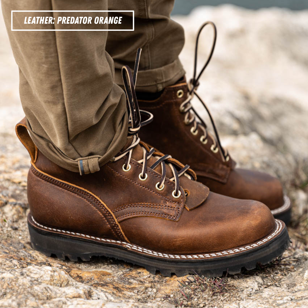 A close-up of a person wearing brown leather lace-up boots with cuffed khaki pants, standing on a rocky outdoor surface. A text label in the top left reads 'LEATHER - PREDATOR ORANGE'.