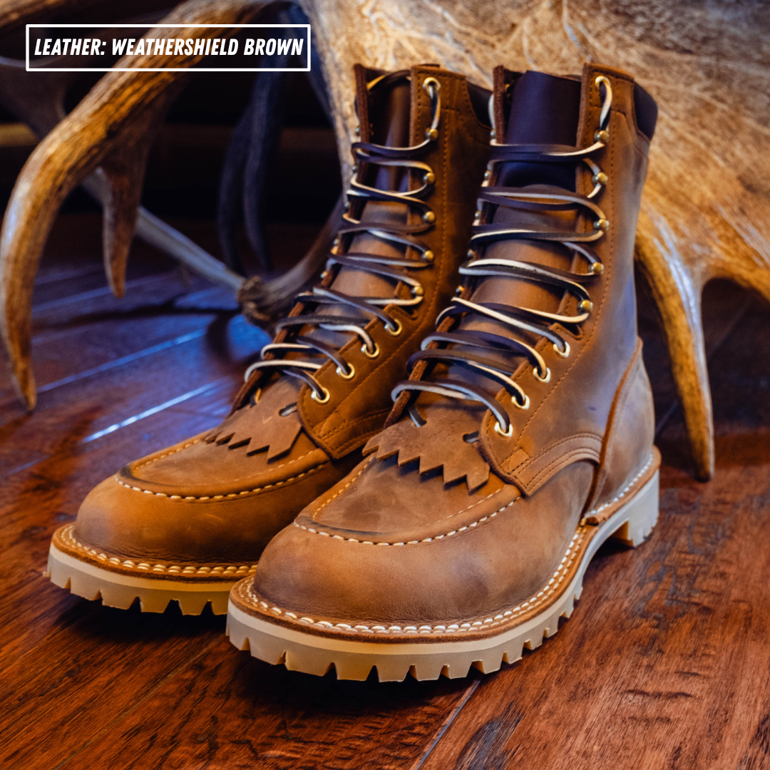 A pair of brown leather lace-up boots with fringed detailing on the vamp, displayed on a wooden surface with antlers in the background. A text overlay in the top-left corner reads 'LEATHER: WEATHERSHIELD BROWN'.