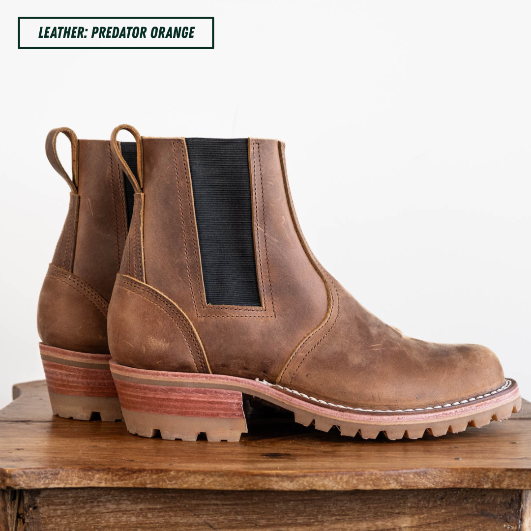 A pair of brown leather Chelsea boots with black elastic side panels and chunky stacked lug soles, displayed on a rustic wooden surface against a white background. A text label in the top left reads 'LEATHER: PREDATOR ORANGE'.