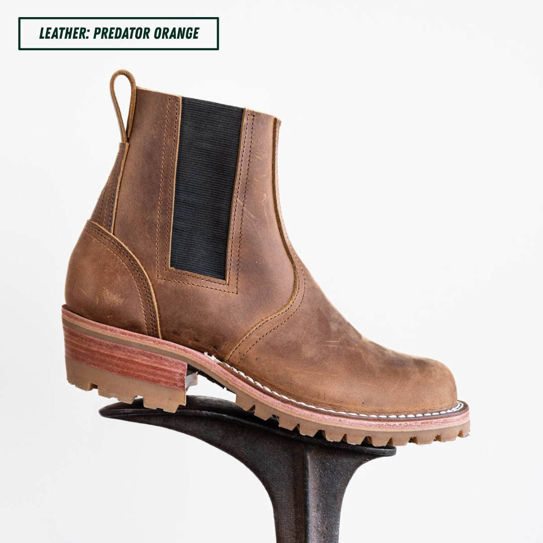 A brown leather Chelsea-style boot with a black elastic side panel, shown in profile on a display stand against a light gray background. A text label at the top reads 'LEATHER: PREDATOR ORANGE'.