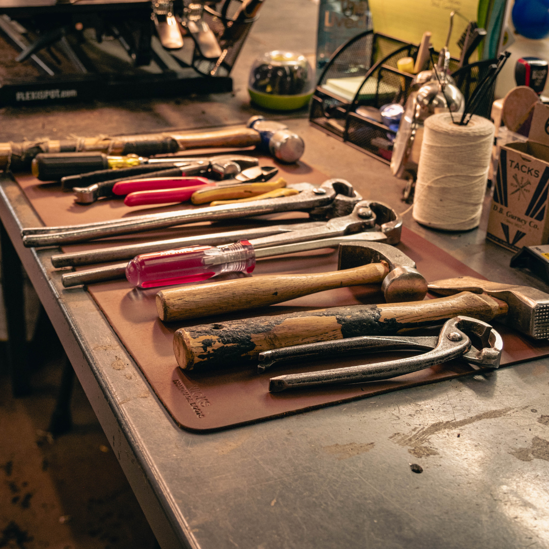 A collection of hand tools, including hammers, pliers, and screwdrivers, laid out on a brown leather work mat on a metal workbench.