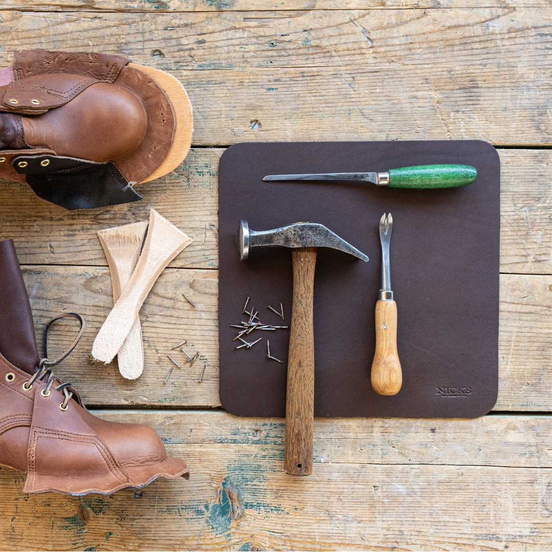 A dark brown leather shoe repair mat laid on a rustic wooden table, surrounded by leatherworking tools including a claw hammer, a green-handled knife, a chisel, nails, and brown leather pieces.