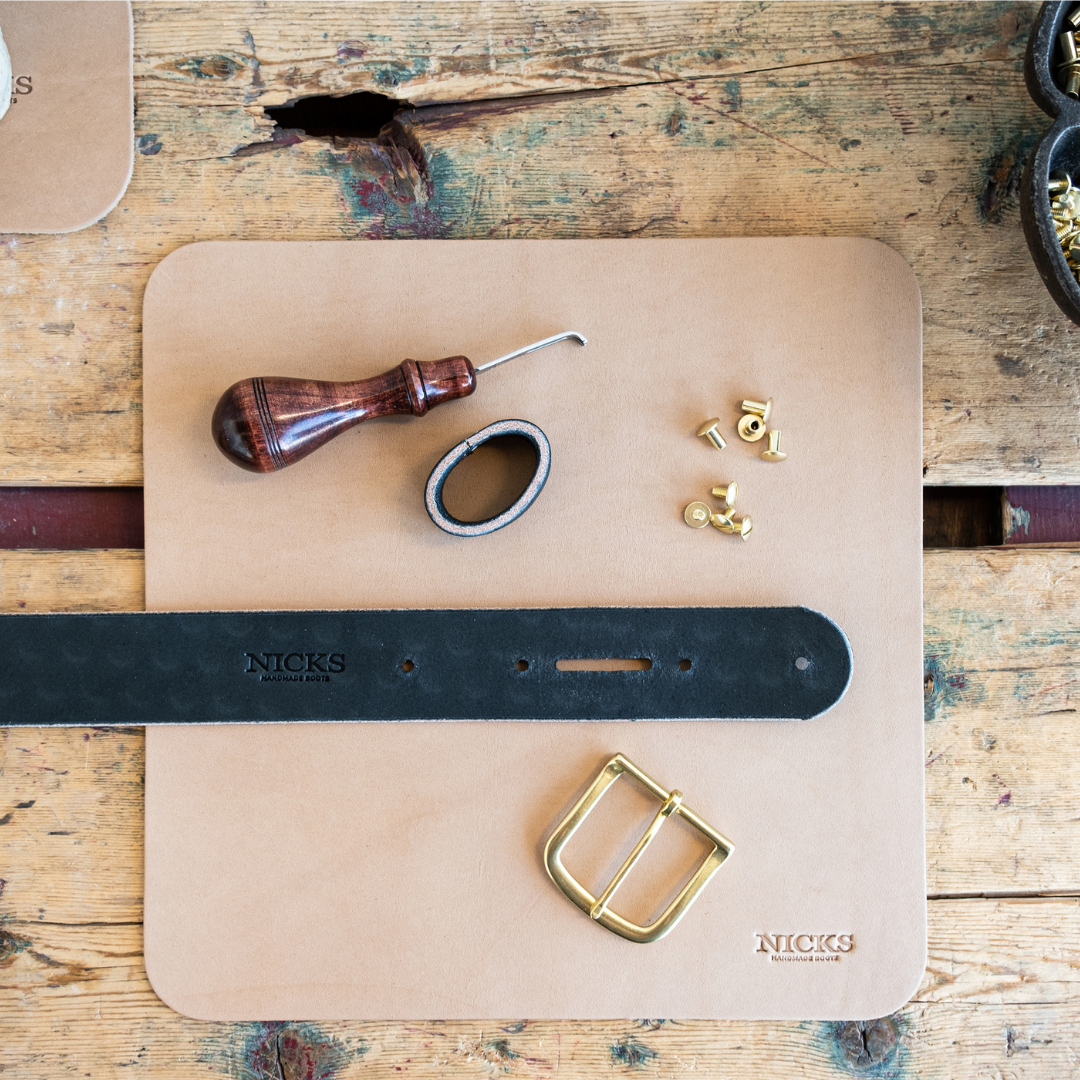 A leather belt and leather working tools arranged on a square tan leather mat over a rustic wooden table. The belt is black with a brass buckle, and the pad shows the NICKS brand name.