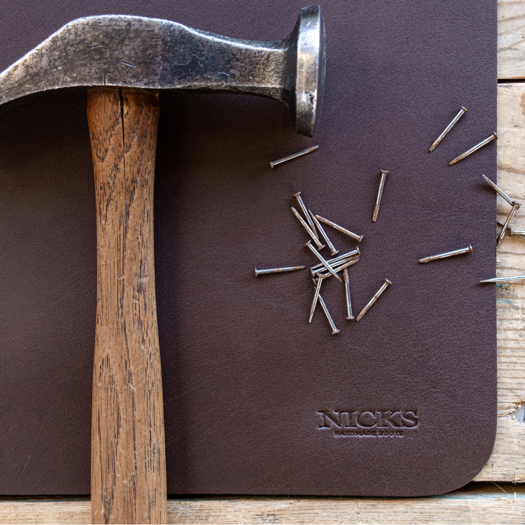 A close-up of a dark brown leather pad with the brand name 'NICKS' embossed in the lower right corner, partially covered by a wooden-handled claw hammer and scattered metal nails on a rustic wood surface.