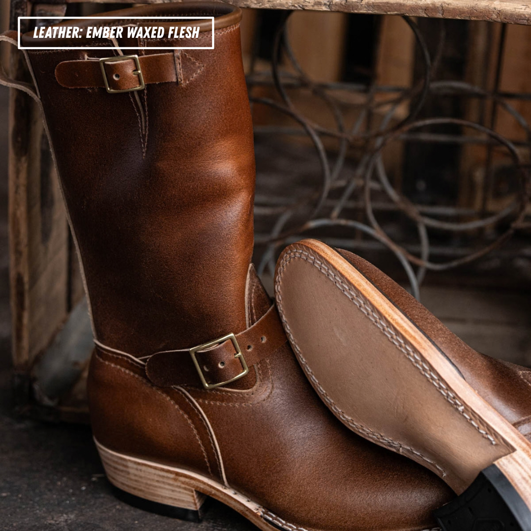 A pair of mid-calf western boots made of rich brown leather, featuring a buckle strap around the calf and a stacked wood-look heel. One boot is standing upright while the other lies on its side to show the tan suede-like sole.