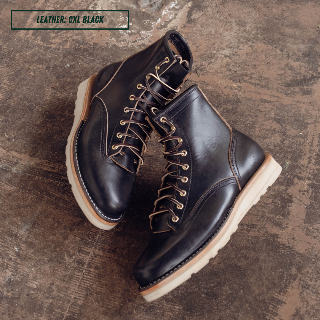 A pair of black leather lace-up ankle boots with tan laces and off-white soles, photographed on a textured brown surface. A text label in the top left reads 'LEATHER: CXL BLACK'.