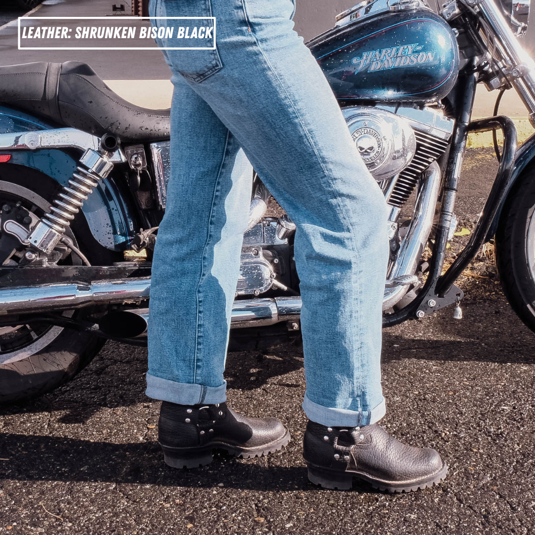 A person wearing cuffed light blue jeans and black leather motorcycle boots stands beside a blue Harley-Davidson motorcycle on asphalt. A label in the top-left corner reads 'LEATHER: SHRUNKEN BISON BLACK'.