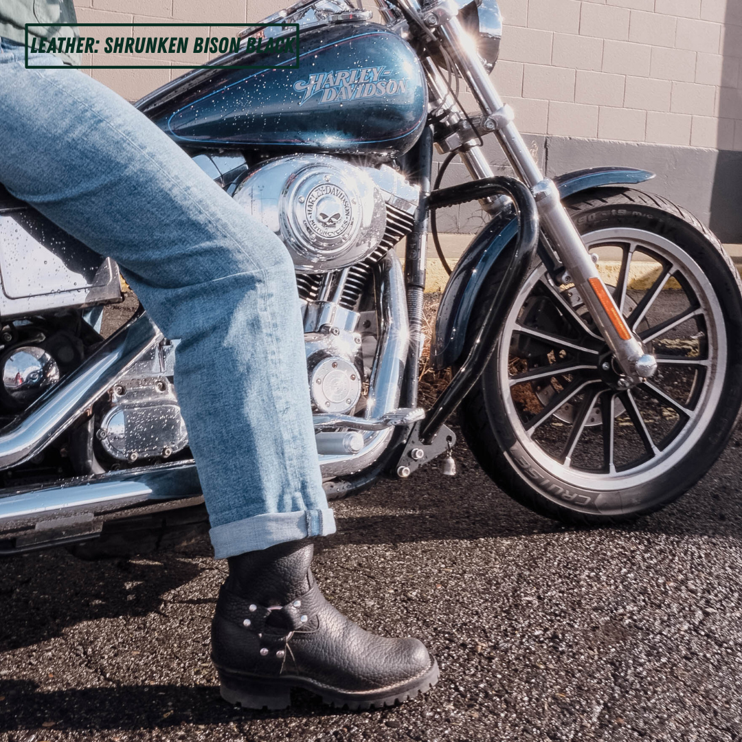 A person wearing black leather studded boot sits on a Harley-Davidson motorcycle outdoors. The photo is cropped to show the person's leg and the boot, with the motorcycle and asphalt pavement visible in the background.