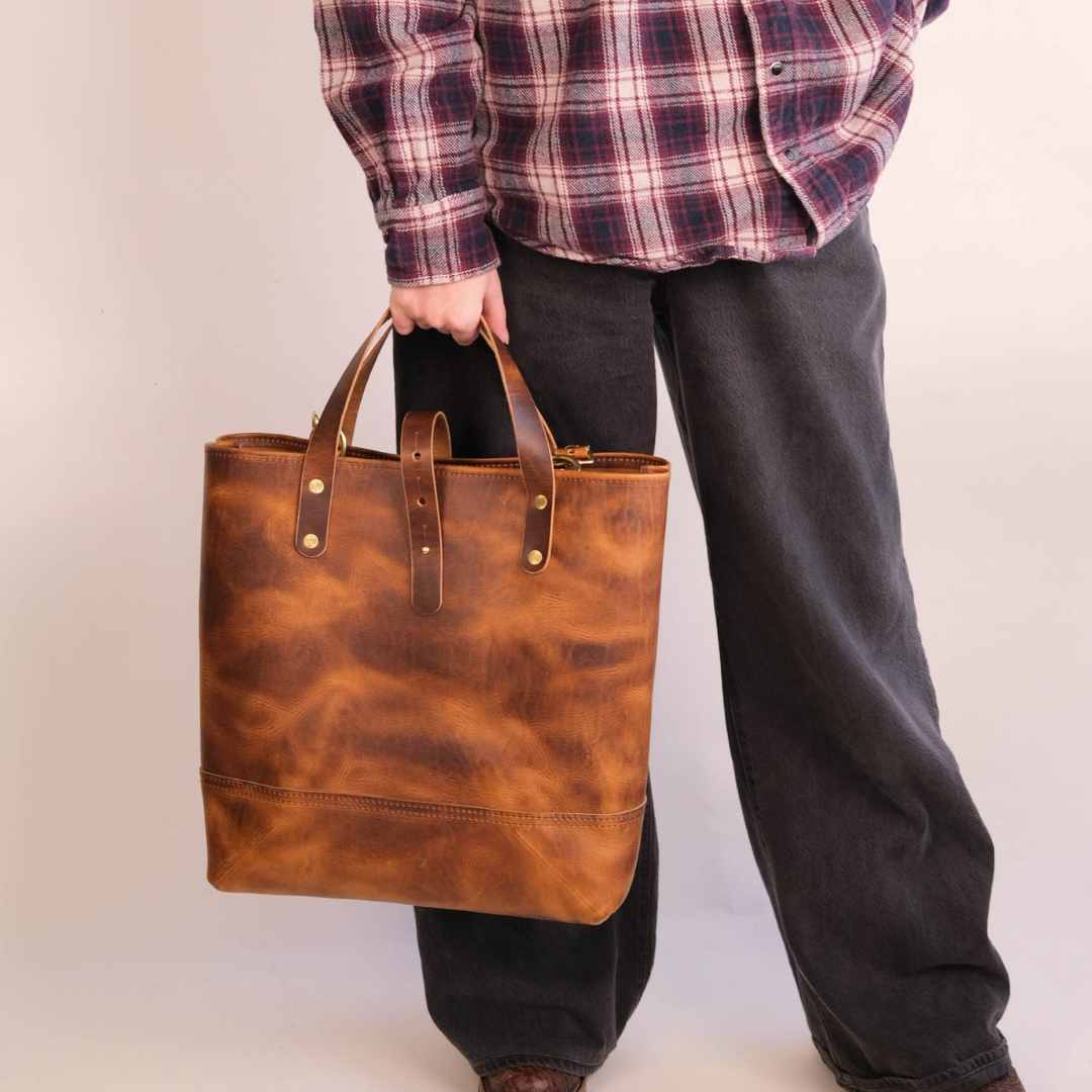 A person wearing a red and navy plaid flannel shirt and dark gray jeans holds a large, distressed brown leather tote bag with brown leather handles and brass hardware against a plain off-white background.