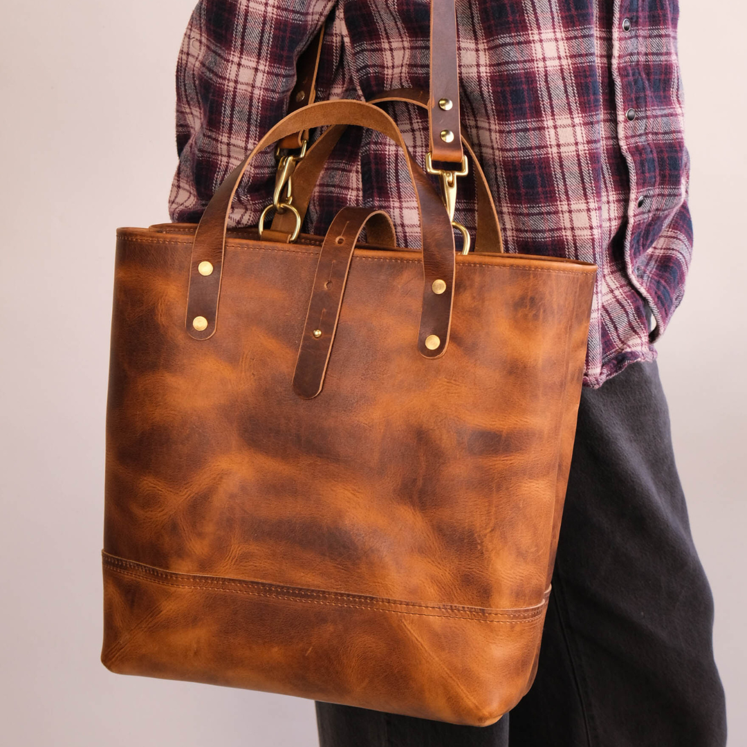 A person wearing a red and navy plaid shirt carries a large, rustic brown leather tote bag with gold-tone hardware against a plain light gray background.