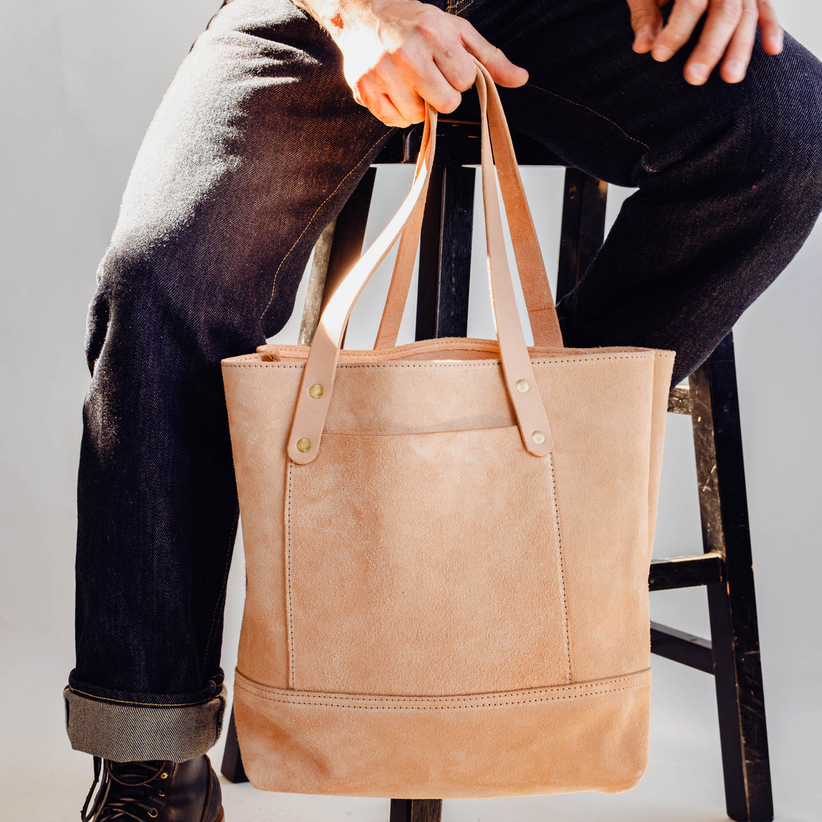 Model holding Veg Tan Suede Tote in front of white background and sitting on wooden stool.