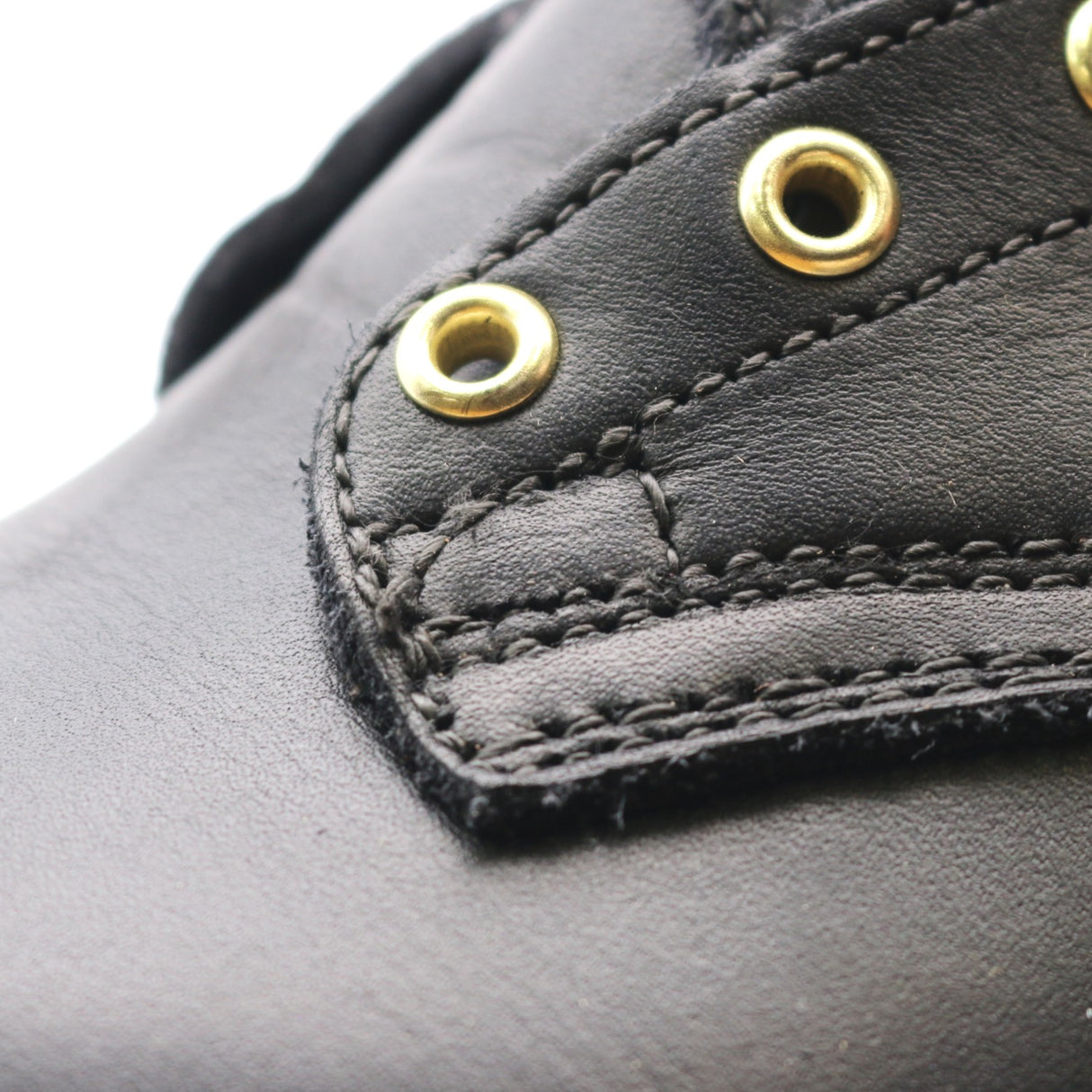 Close-up of a black leather shoe upper with gold-tone metal eyelets and detailed stitching.