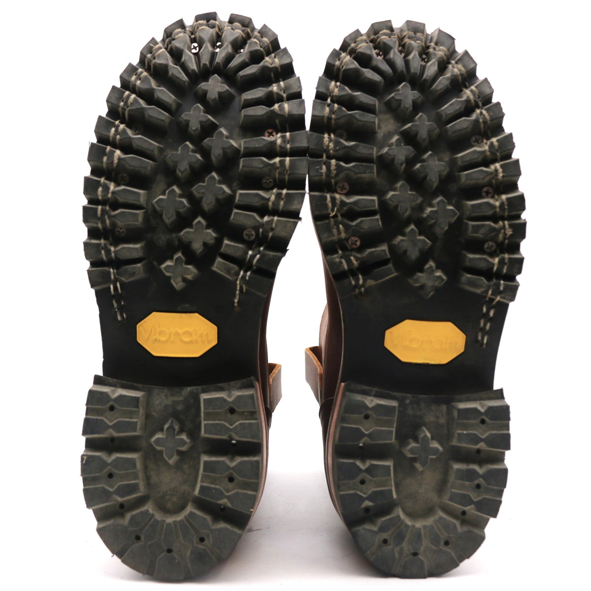 The soles of a pair of brown boots, showing deep lugs, Vibram branding in yellow on the heel, and a rugged black tread pattern on a white background.
