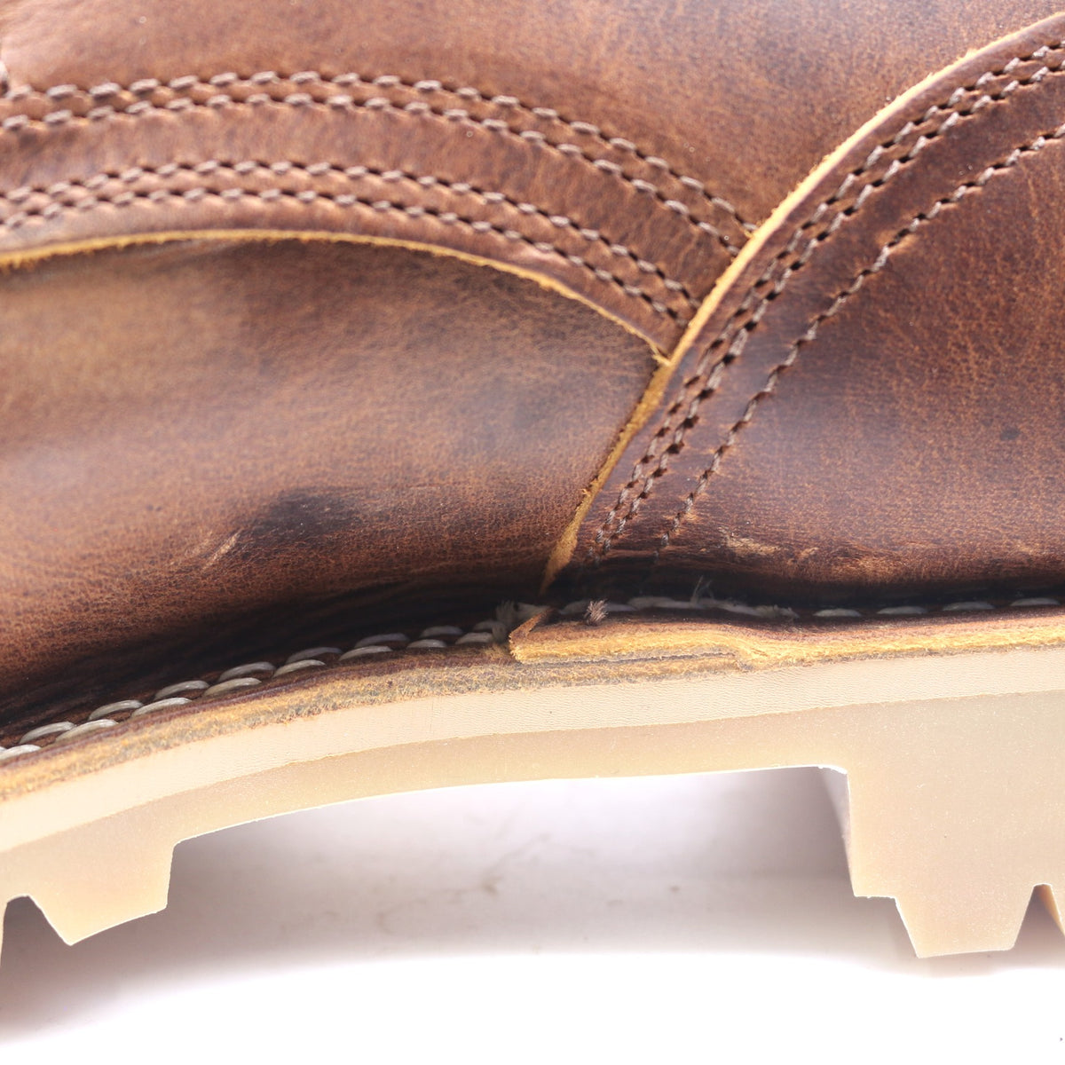 Close-up of a brown leather boot showing the textured leather upper, contrast stitching, and a light beige sole edge against a white background.