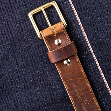 A close-up of a brown leather watch strap with a brass buckle, laid diagonally on dark navy denim fabric with red and white contrast stitching.
