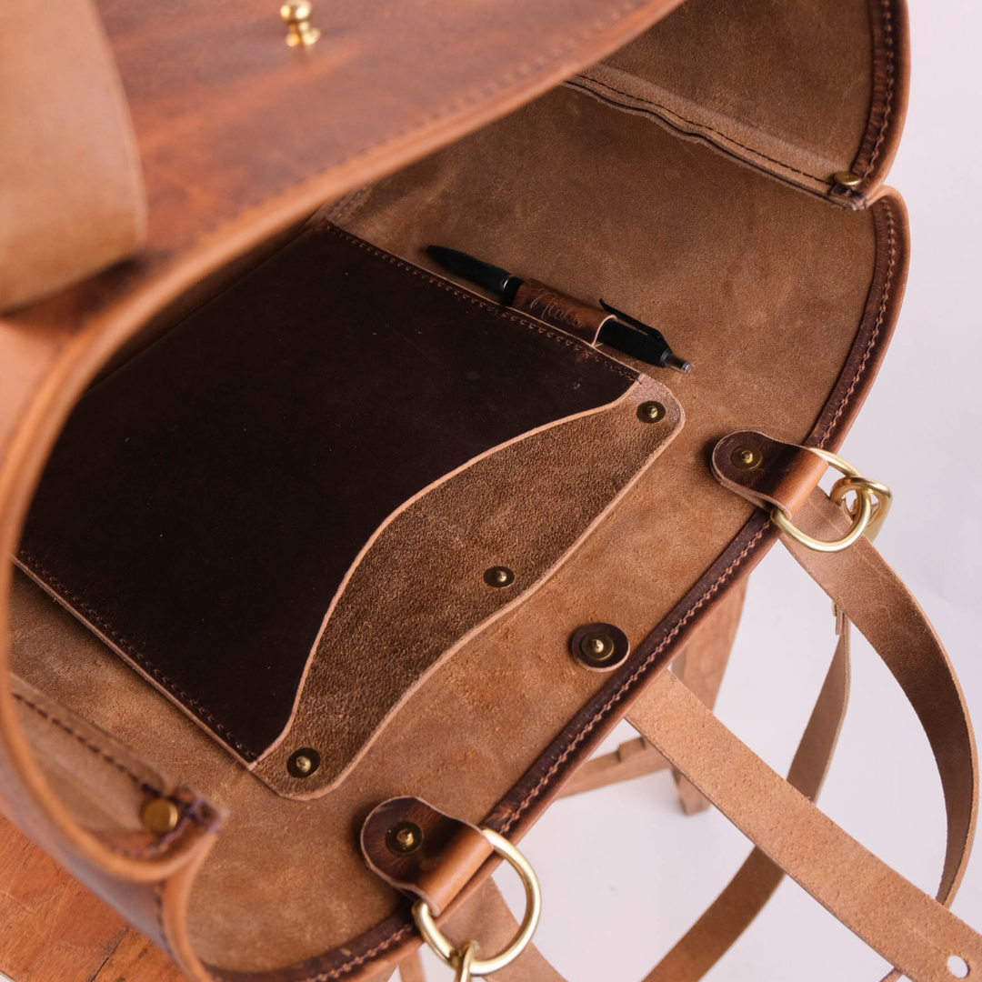 Close-up of an open brown leather bag showing the interior lining, pen pocket with a black pen, brass-tone hardware, and a strap against a light neutral background.