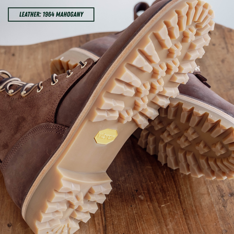 A close-up of a brown leather lace-up boot with a chunky beige lug sole, shown on a wooden surface. A text label in the top left reads 'LEATHER: 1964 MAHOGANY'.
