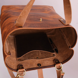A close-up of an open brown leather bag showing its interior, including a dark insert pocket, a pen loop, brass-tone hardware, and a strap hanging down against a plain light background.