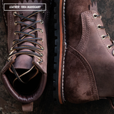 Two brown leather lace-up boots shown in a close-up product shot on a dark textured surface. One boot is angled to show the lace area and opening, and the other shows the side profile with stitching and sole.