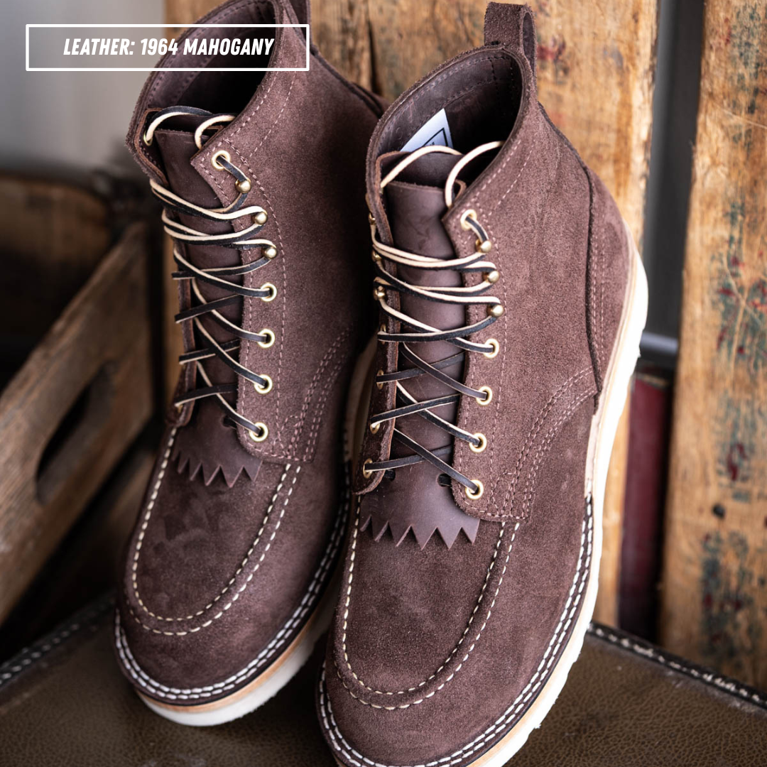 A pair of dark brown suede lace-up boots with white contrast stitching and metal eyelets, displayed on a leather surface with rustic wooden crates in the background. A text overlay in the top left reads 'LEATHER: 1964 MAHOGANY'.