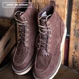 A pair of dark brown suede lace-up boots with white contrast stitching and metal eyelets, displayed on a leather surface with rustic wooden crates in the background. A text overlay in the top left reads 'LEATHER: 1964 MAHOGANY'.
