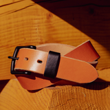A tan leather belt with a black buckle and black keeper loop, coiled on a rustic wooden surface.