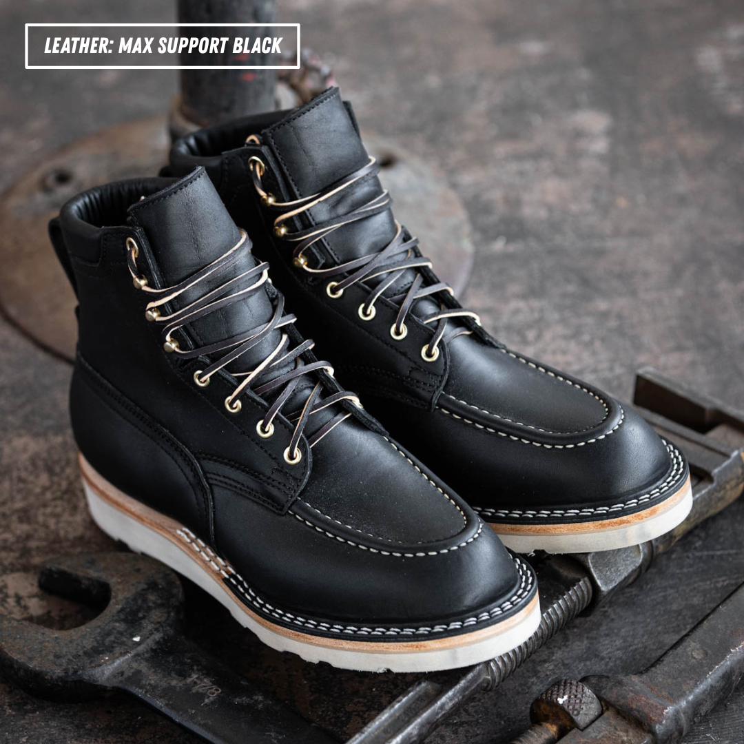 A pair of black leather lace-up work boots with white stitching and white soles, displayed on a rustic metal surface. A text overlay in the top-left corner reads 'LEATHER: MAX SUPPORT BLACK'.