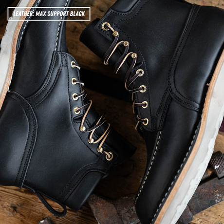 A pair of black leather lace-up boots with gold-toned metal eyelets and white stitching, shown on a rustic wooden surface. A text overlay in the top-left corner reads 'LEATHER: MAX SUPPORT BLACK'.