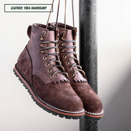 A pair of brown suede lace-up boots hanging from straps next to a dark vertical pole, with a text label in the top-left corner reading 'LEATHER: 1964 MAHOGANY'.