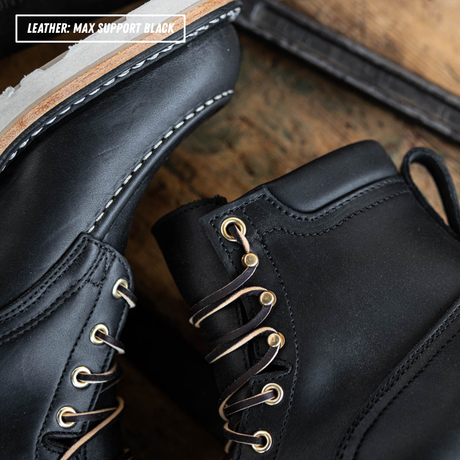 Close-up of black leather lace-up boots with gold-tone eyelets and laces, shown on a rustic wooden surface. A text overlay in the top left reads 'LEATHER: MAX SUPPORT BLACK'.