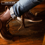 Model wearing brown Moctoepro while sitting in chair. Label on top left corner that reads "LEATHER: 1964 BROWN."