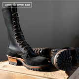 A pair of tall black leather lace-up boots with chunky lug soles and stacked heels, displayed on a wooden surface against a dark gray background. One boot stands upright while the other lies on its side showing the sole.
